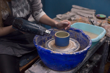 Fototapeta na wymiar Girl with an industrial hairdryer dries clay cup on potter's wheel. Pottery hobby. Handcrafted earthenware