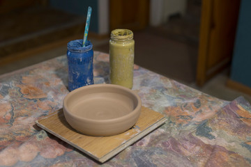 Unpainted clay plate on the table next to jars with paints. Beige plate earthenware standing on table in pottery workshop