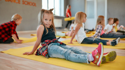 Waiting for teacher. Portrait of a pretty little girl sitting on the yellow yoga mat and looking at camera while having a yoga class in the dance studio. Children doing gymnastic exercises