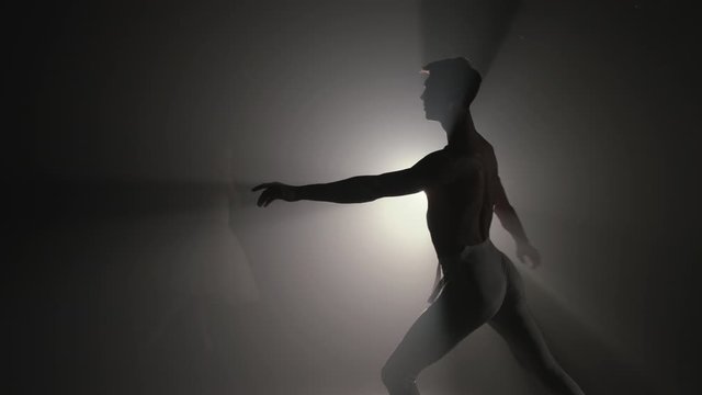 Professional choreographer is working on creating performance. Young handsome male dancer practicing in classical ballet in studio with smoke back light. Man in white tights. 