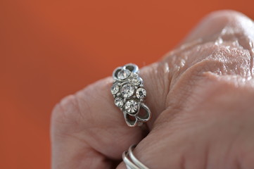 A diamond ring on the finger of an old woman in front of an orange background.