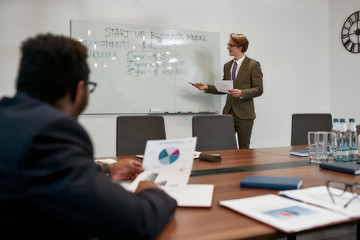 Discussing income. Portrait of young and smart businessman in classic wear holding financial report and pointing at white blackboard while explaining something to his colleagues