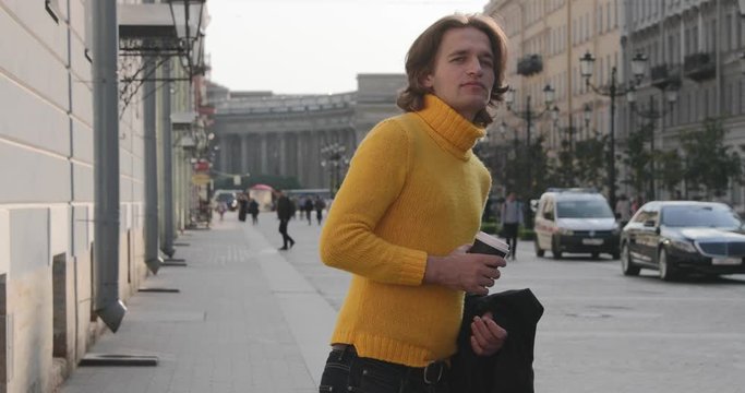 The handsome men is waiting someone, drinks coffee, he dressed in a yellow sweater and black raincoat, Bolshaya Konyushennaya street and Kazan Cathedral on background, sunny day