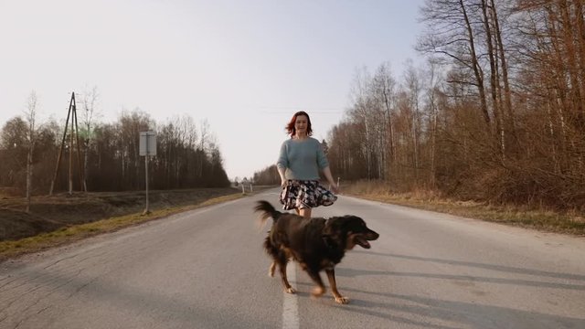 Cheerful redhead woman runs with her big dog towards you during sunset in middle of road. Slow motion