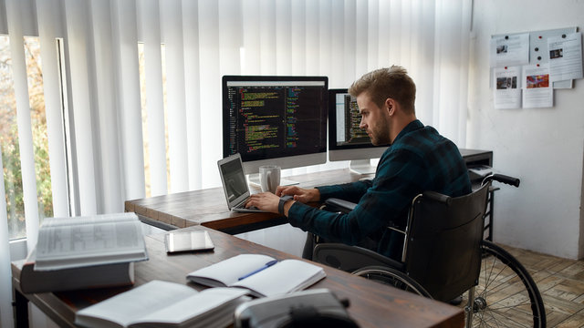 IT professional. Young caucasian male web developer in a wheelchair writing program code while sitting at workplace with three monitors in the modern office. HTML, PHP and javascript