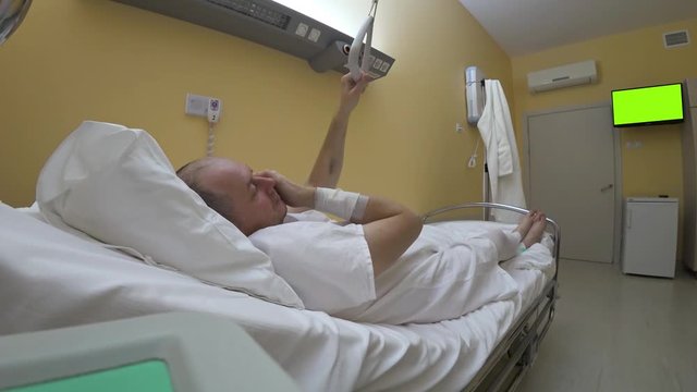 A recovering patient talks on the phone lying on a bed in a hospital room against the background of the green screen tv chromakey