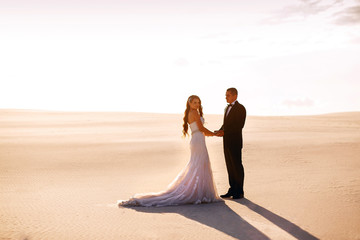 Fototapeta na wymiar Bride and groom holding hands in the sand desert at sunset