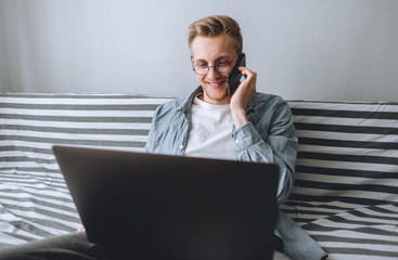 Smilling young man works at home with a laptop and talking on a cell phone. Remote work, home education