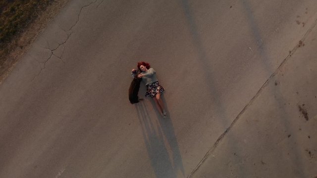 Aerial view: young redhead woman lays on asphalt road and plays with pariah dog during sunset rays