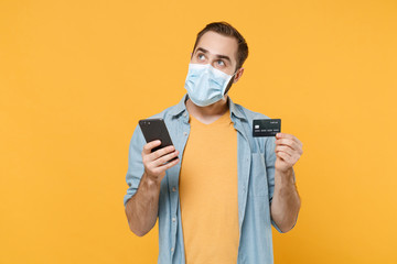 Pensive young man in sterile face mask posing isolated on yellow background studio. Epidemic pandemic coronavirus 2019-ncov sars covid-19 flu virus concept. Using mobile phone, hold credit bank card.