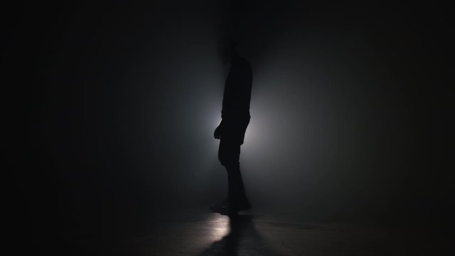 Silhouette of young man covers his face with hands from fatal disappointment, failure. Depressed lost man in smoky dark studio. Drama, emotions concept