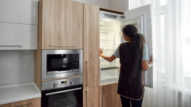 Making your life easier. Young afro american house maid in uniform cleaning refrigerator in the modern kitchen. Cleaning services