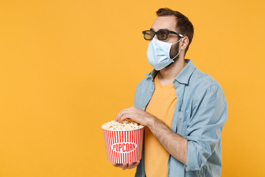 Shocked young man in 3d glasses face mask isolated on yellow background. Epidemic pandemic coronavirus 2019-ncov sars covid-19 flu virus concept. Watching movie film hold bucket popcorn looking aside.