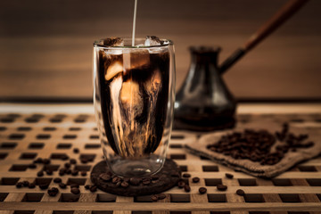 Adding milk to a big glass of iced coffee, coffee beans around, cezve. Wooden table and wooden background. Horizontal photo.