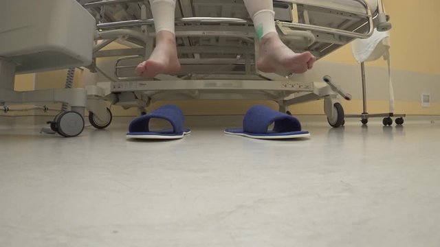 Close-up, bottom view, in a hospital room, the patient sits on a mechanical electrified bed. In front of him are blue slippers. The bed slowly lowers, the legs slip into the slippers and leave.