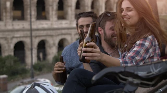 Three young friends tourists with bikes sitting on bench in front of colosseum under tree at sunset drinking beers having fun talking laughing chilling in Rome slow motion