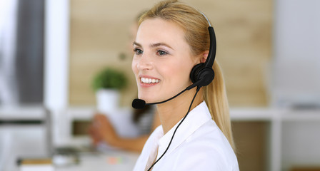Blonde business woman using headset for communication and consulting people at customer service office. Call center. Group of operators at work at the background