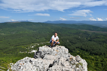 The man squatted on the edge of the cliff, behind him a beautiful view of nature, green forests, a blue sky with white clouds, a picturesque nature panorama.