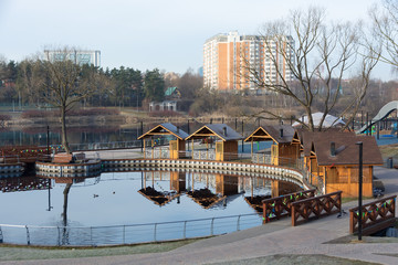 Fototapeta na wymiar Panorama of small city park with pond, line of pavilions and bridge. Early spring morning