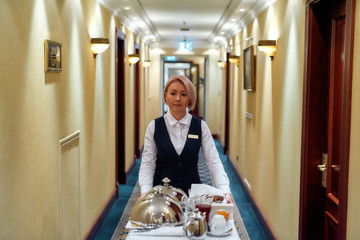 Experience the passion of hospitality. Waitress in uniform walking along the hall while delivering tray with food in a room of hotel. Room service. - obrazy, fototapety, plakaty