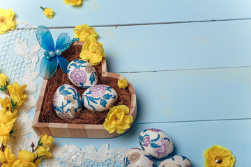 Decorated Easter eggs in heart shaped box. White lace, yellow artificial flowers, dragonfly toy on blue wooden background. Copy space.