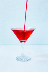 Pouring red syrup to a martini cocktail glass with a drink, light blue background, natural light vertical photo