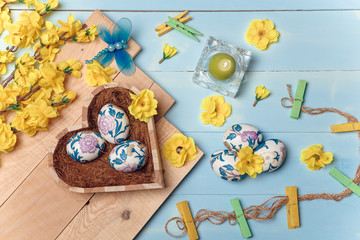 Heart shaped box with decorated Easter eggs, yellow flowers, burning candle and brown boards on wooden blue background. Spring and Easter concept. Top view, flatlay, copy space.