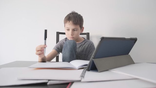 Sad child sitting at desk with tablet computer and many books. Boy feels bored, because he do not unterstand homework, he play with pen. Education, learning difficulties, online learning concept