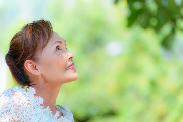 Closeup side face of a healthy Asian middle-aged woman dress in Thai style clothes, Portrait old lady resting outdoors looking up smiling happy in the park, copy space on green tree nature background