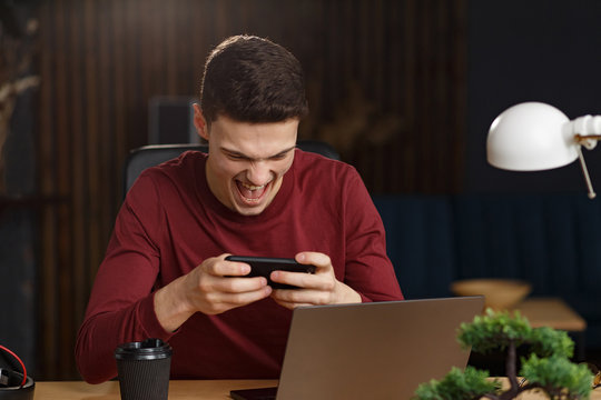 Young Man Playing Games On Mobile Phone In Home Office With Laptop. Relaxation, Taking A Break. Young People Working With Mobile Devices.