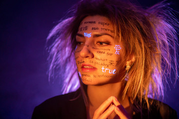 Girl with Disheveled Hair in Dramatic Purple light with written different language words on her face. Beautiful model with UV Fluorescent neon creative make-up. face close up
