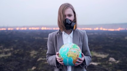 A student girl in a protective mask or a respirator holds a globe in her hands. on the background of earth in smoke and fire. burning grass leaves emissions Earth conservation concept, disaster