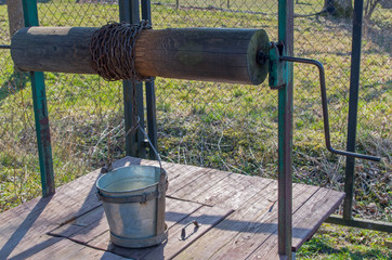 Standing on closed wooden door of well metal galvanized bucket attached to  hoops frame and fixed by handle to chain wound on log wheel and axle, which held on green iron poles, on background of fence © ANDY RELY
