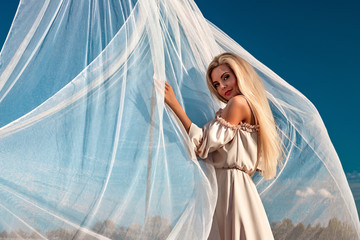 Portrait of a beautiful girl, blonde with long hair in a field, on a background of white transparent fabric