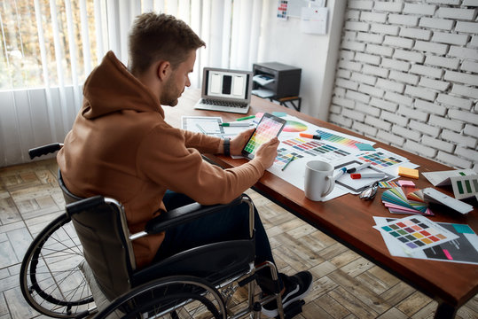 Doing creative work. Young male UX UI designer in a wheelchair choosing color samples for new project while working at his creative workplace in the modern office