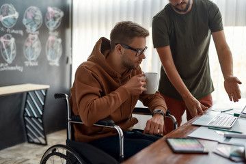 Working on new project. Young concentrated male designer in a wheelchair drinking coffee and discussing some ideas with his colleague while working together in the creative office
