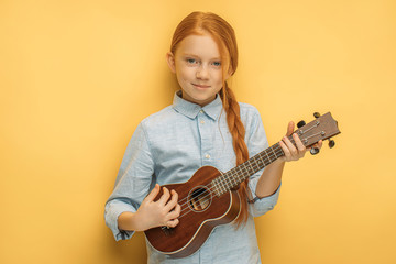 portrait of awesome caucasian child girl playing ukulele, attractive girl with natural red hair keen on music and instruments. isolated yellow background - obrazy, fototapety, plakaty