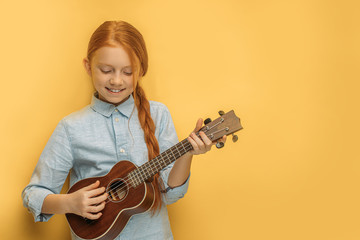 Fototapeta premium beautiful caucasian child girl playing ukulele, little girl with natural red hair and freckles loves music and instruments. isolated yellow background