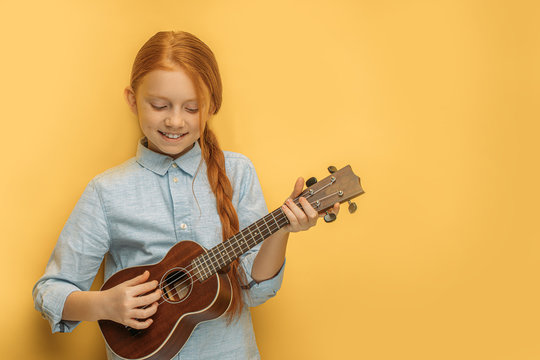 beautiful caucasian child girl playing ukulele, little girl with natural red hair and freckles loves music and instruments. isolated yellow background