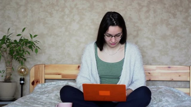 Young attractive girl is actively browsing the news feed on a laptop while sitting on a bed at home. Young Cute girl is working on a laptop while sitting on a bed.