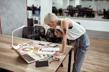 New project. Young stylish blonde tattooed female designer making some sketches and working with color swatch samples while standing near office desk