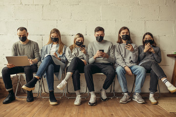 young caucasian people sit together in protective masks, they protect themselves from coronavirus, covid-19 disease. sit using laptop and smartphone. danegrous infection, virus, global problem.