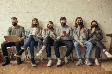 Fototapeta premium young caucasian people sit together in protective masks, they protect themselves from coronavirus, covid-19 disease. sit using laptop and smartphone. danegrous infection, virus, global problem.