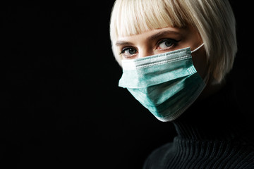 Woman wearing medical protective face mask looks at camera, posing on black background. Copy, empty...
