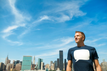 Fototapeta na wymiar Smiling young man in a T-shirt with a blank speech bubble standing in front of the city skyline under bright blue sky