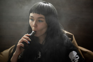 Obraz premium Vaping teenager. Young pretty white caucasian teenage brunette girl with blue eyes and problem skin smoking an electronic cigarette indoors. Deadly bad habit. Vape activity.