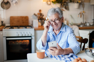 Senior woman using mobile phone at home kitchen. Happy retired person shopping online. People connection, virtual communication, distance healthcare. Pensioner in social distancing, isolation.