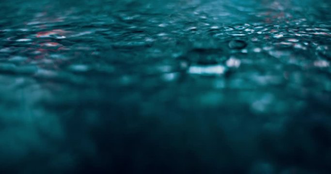 Slow motion rain drops falling into puddle on asphalt at night. Big modern city lights mirror reflection on water surface. Close-up of raining outside in wet season. Raindrops falling down
