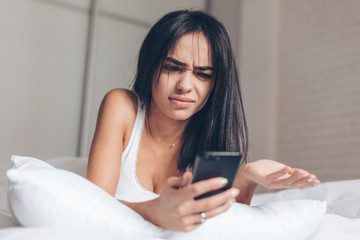 Obraz na płótnie Canvas Young Annoyed angry woman looking on mobile phone in bed. Smiling Brunette Girl holding mobile phone