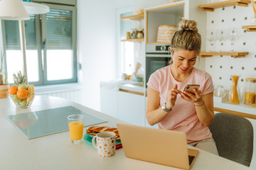 Naklejka premium Portrait of young woman with messy blonde hair looking at her smartphone while sitting in front of laptop at home in kitchen. Young woman is working from home and having breakfast at same time.